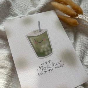 Puede incluir: Tarjeta de felicitaci&oacute;n de acuarela con un dibujo de una bebida de matcha sonriente con una pajita. La tarjeta dice "have so Matcha fun on your birthday" en escritura negra. Flores secas de color marr&oacute;n claro en el fondo.