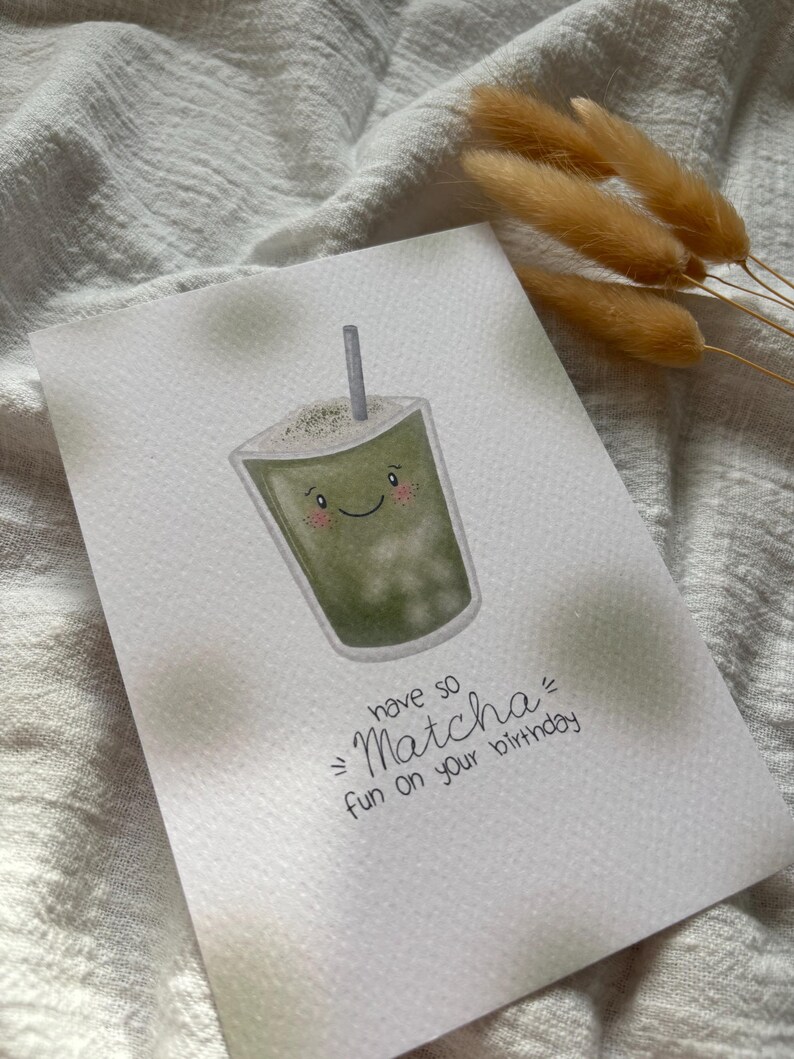 Puede incluir: Tarjeta de felicitaci&oacute;n de acuarela con un dibujo de una bebida de matcha sonriente con una pajita. La tarjeta dice "have so Matcha fun on your birthday" en escritura negra. Flores secas de color marr&oacute;n claro en el fondo.