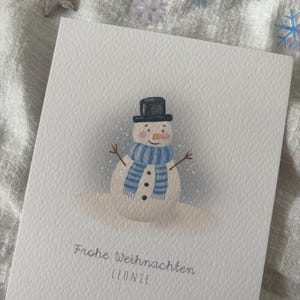 May include: A white greeting card featuring a watercolor illustration of a snowman wearing a black top hat and a blue striped scarf. The card has the text "Frohe Weihnachten LEONIE" in a light gray font.