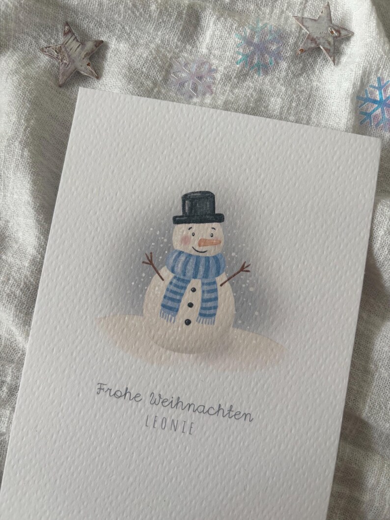 May include: A white greeting card featuring a watercolor illustration of a snowman wearing a black top hat and a blue striped scarf. The card has the text "Frohe Weihnachten LEONIE" in a light gray font.