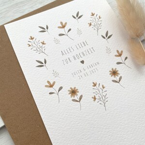 May include: A white card with a brown envelope. The card has a floral design with brown and yellow flowers and leaves. The text on the card reads "Alles Liebe zur Hochzeit" and "Julia & Fabian 24.08.2023".