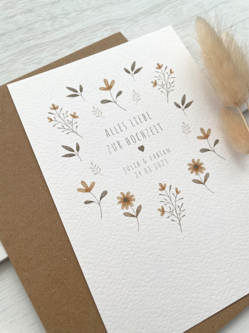 May include: A white card with a brown envelope. The card has a floral design with brown and yellow flowers and leaves. The text on the card reads "Alles Liebe zur Hochzeit" and "Julia & Fabian 24.08.2023".