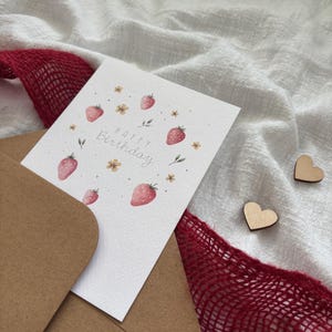 May include: A white card with a brown envelope. The card has a watercolor illustration of strawberries and flowers. The text "Happy Birthday" is written in cursive on the card.