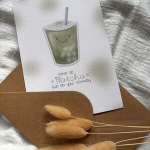 Puede incluir: Una tarjeta de felicitaci&oacute;n con una ilustraci&oacute;n de bebida de matcha y el texto "have so Matcha fun on your birthday". La tarjeta est&aacute; dentro de un sobre marr&oacute;n, con hierba de cola de conejo seca en frente. La bebida de matcha tiene una cara sonriente y una pajita.