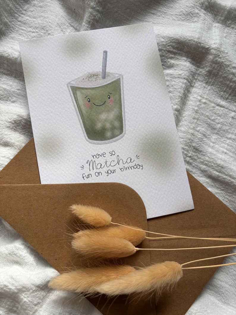 Puede incluir: Una tarjeta de felicitaci&oacute;n con una ilustraci&oacute;n de bebida de matcha y el texto "have so Matcha fun on your birthday". La tarjeta est&aacute; dentro de un sobre marr&oacute;n, con hierba de cola de conejo seca en frente. La bebida de matcha tiene una cara sonriente y una pajita.