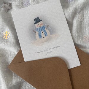 May include: A Christmas card featuring a snowman illustration with a black top hat and blue scarf. The card is inside a brown envelope, set against a white, textured background with decorative snowflakes and stars.