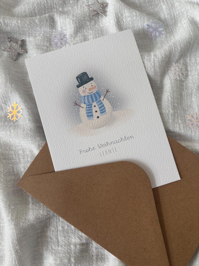 May include: A Christmas card featuring a snowman illustration with a black top hat and blue scarf. The card is inside a brown envelope, set against a white, textured background with decorative snowflakes and stars.
