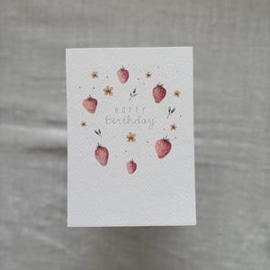 May include: A white card with a watercolor illustration of strawberries and flowers. The text "Happy Birthday" is written in cursive.