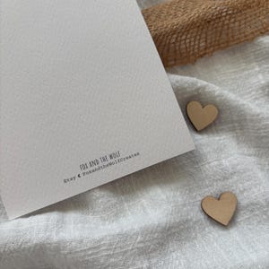 May include: A white card with the text "Fox and the Wolf" and "Etsy Foxandthewolfcreates" printed on it. Two small wooden hearts are on a white fabric surface.