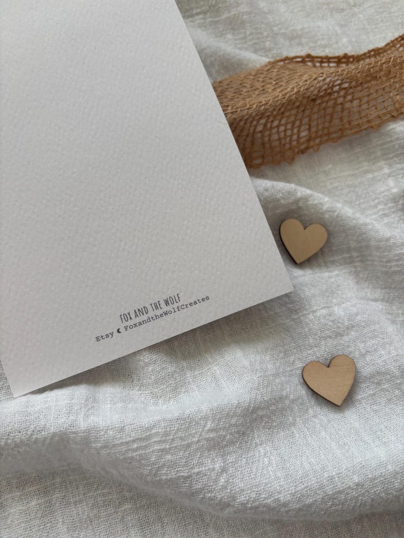 May include: A white card with the text "Fox and the Wolf" and "Etsy Foxandthewolfcreates" printed on it. Two small wooden hearts are on a white fabric surface.