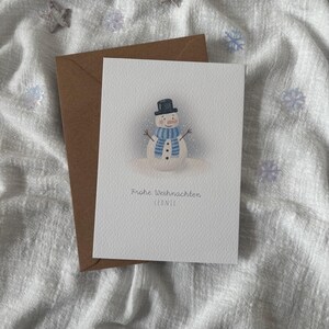 May include: A Christmas card featuring a watercolor snowman wearing a black top hat and a blue striped scarf. The card is paired with a brown envelope. The text "Frohe Weihnachten Leonie" is printed on the card.