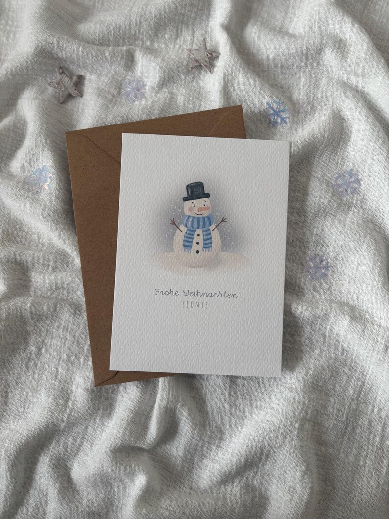 May include: A Christmas card featuring a watercolor snowman wearing a black top hat and a blue striped scarf. The card is paired with a brown envelope. The text "Frohe Weihnachten Leonie" is printed on the card.