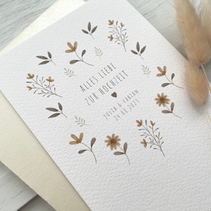 May include: A white card with a floral design and the text "Alles Liebe zur Hochzeit" in black. Below the text is a small heart and the text "Julia & Fabian 24.08.2023" in black.
