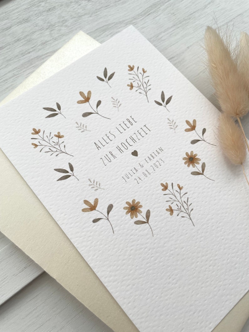 May include: A white card with a floral design and the text "Alles Liebe zur Hochzeit" in black. Below the text is a small heart and the text "Julia & Fabian 24.08.2023" in black.