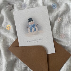 May include: A Christmas card featuring a snowman illustration, wearing a black top hat and a blue scarf, is placed inside a brown envelope. The card reads "Frohe Weihnachten" and the name "LEONIE". Decorative stars and snowflakes are scattered around.