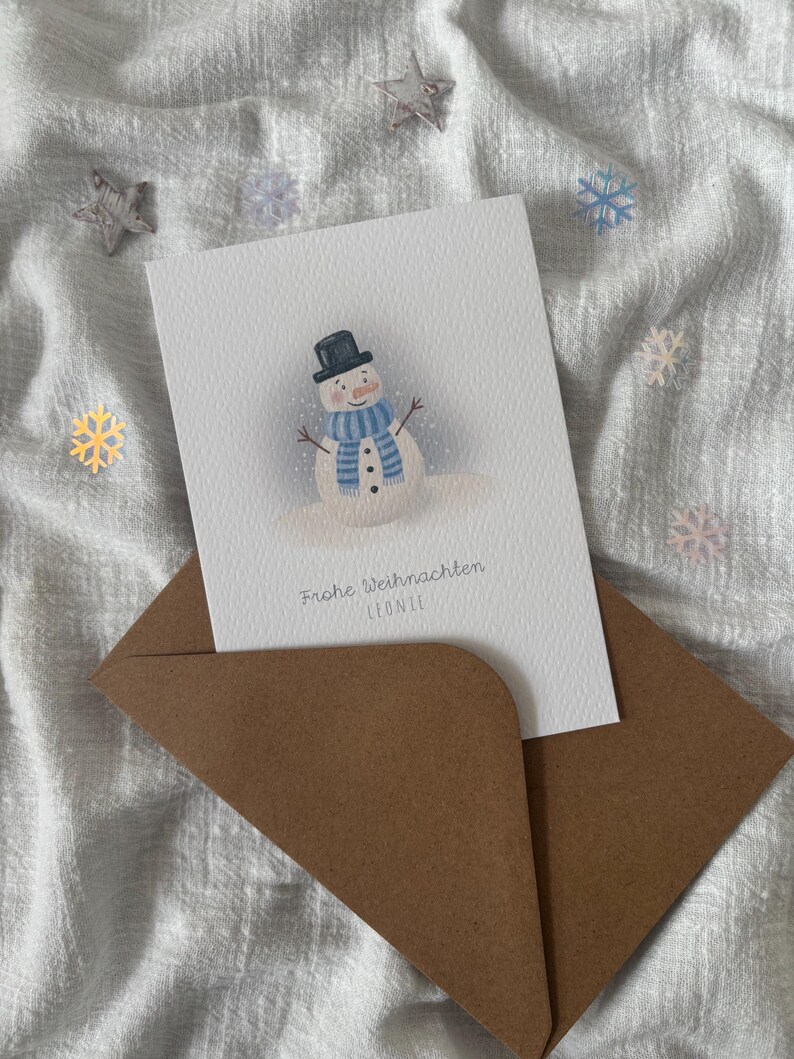May include: A Christmas card featuring a snowman illustration, wearing a black top hat and a blue scarf, is placed inside a brown envelope. The card reads "Frohe Weihnachten" and the name "LEONIE". Decorative stars and snowflakes are scattered around.