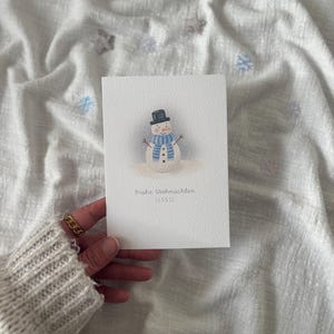 May include: A watercolor Christmas card featuring a snowman with a black top hat, blue scarf, and carrot nose. The card has the text "Frohe Weihnachten" and the name "LEONIE" in a delicate font.