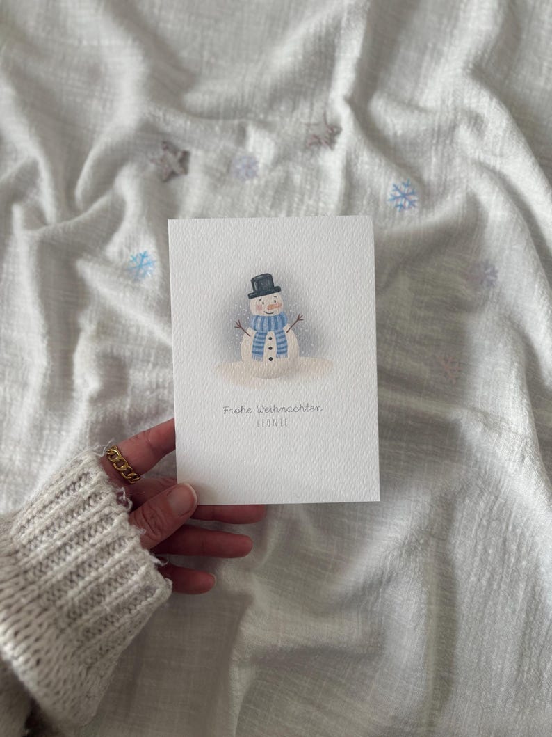 May include: A watercolor Christmas card featuring a snowman with a black top hat, blue scarf, and carrot nose. The card has the text "Frohe Weihnachten" and the name "LEONIE" in a delicate font.