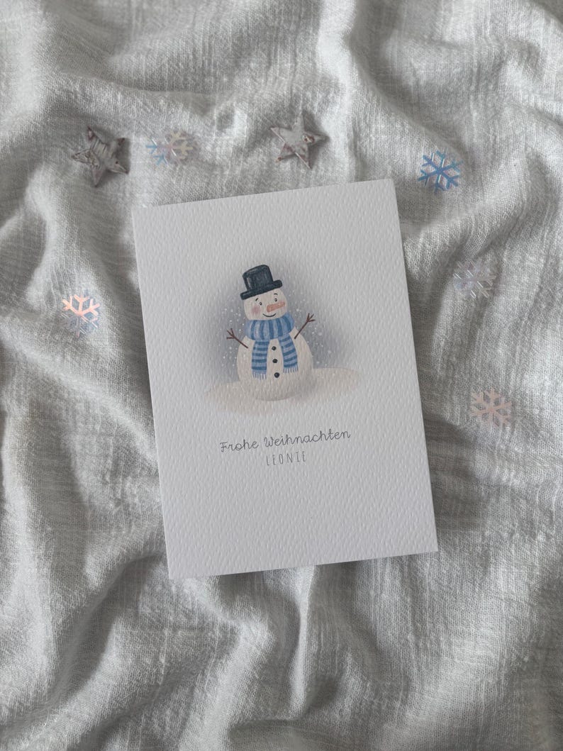 May include: A white greeting card featuring a snowman illustration. The snowman wears a black top hat and a blue and white striped scarf. The card has the text "Frohe Weihnachten LEONIE". Decorative stars and snowflakes are scattered around the card.