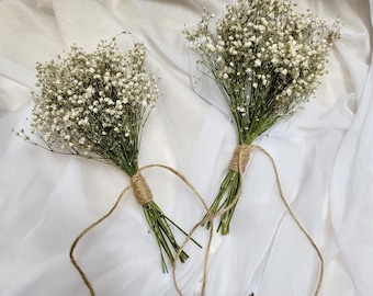 May include: Two small bouquets of white baby's breath flowers tied with twine. The flowers have small white blooms and green stems. The twine is a natural brown color, and the background is a white fabric.