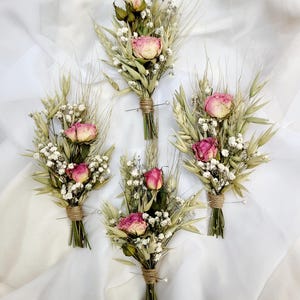May include: Four small bouquets of dried flowers. Each bouquet features a pink rose, white baby's breath, and wheat stalks, tied with twine. The bouquets are arranged on a white, textured fabric.