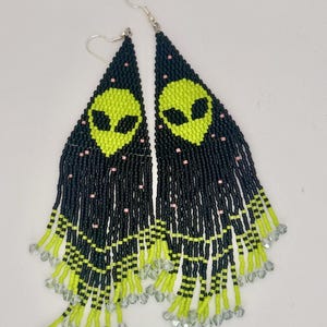 May include: A pair of beaded earrings featuring a neon green alien face design on a black background. The earrings have a triangular shape with long fringe, accented with pink beads and clear crystal beads at the ends.