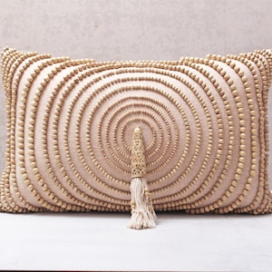 Puede incluir: Una almohada beige con un patr&oacute;n circular de cuentas de madera. La almohada tiene un borla de hilo blanco.