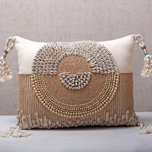 May include: A white throw pillow with a circular design of beige and white beads and shells. The pillow has a fringe trim and tassels on the corners.