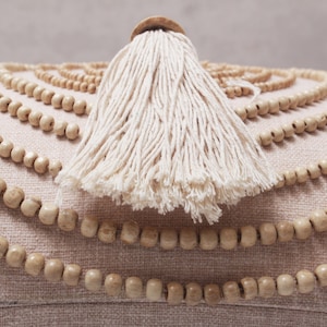 Puede incluir: Un coj&iacute;n redondo beige con un borla blanca y un ribete decorativo de cuentas de madera.