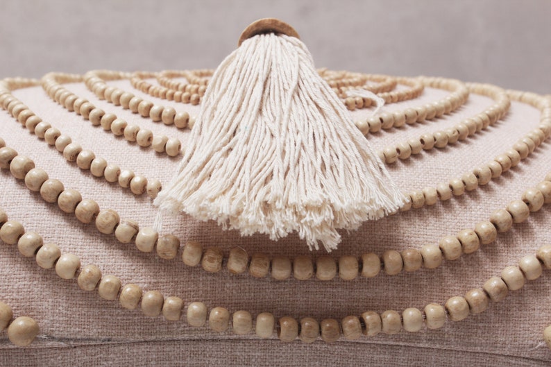 Puede incluir: Un coj&iacute;n redondo beige con un borla blanca y un ribete decorativo de cuentas de madera.
