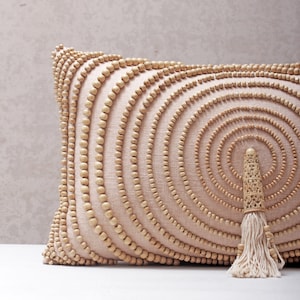Puede incluir: Una almohada beige con un patr&oacute;n circular de cuentas de madera. La almohada tiene un borla de hilo blanco.