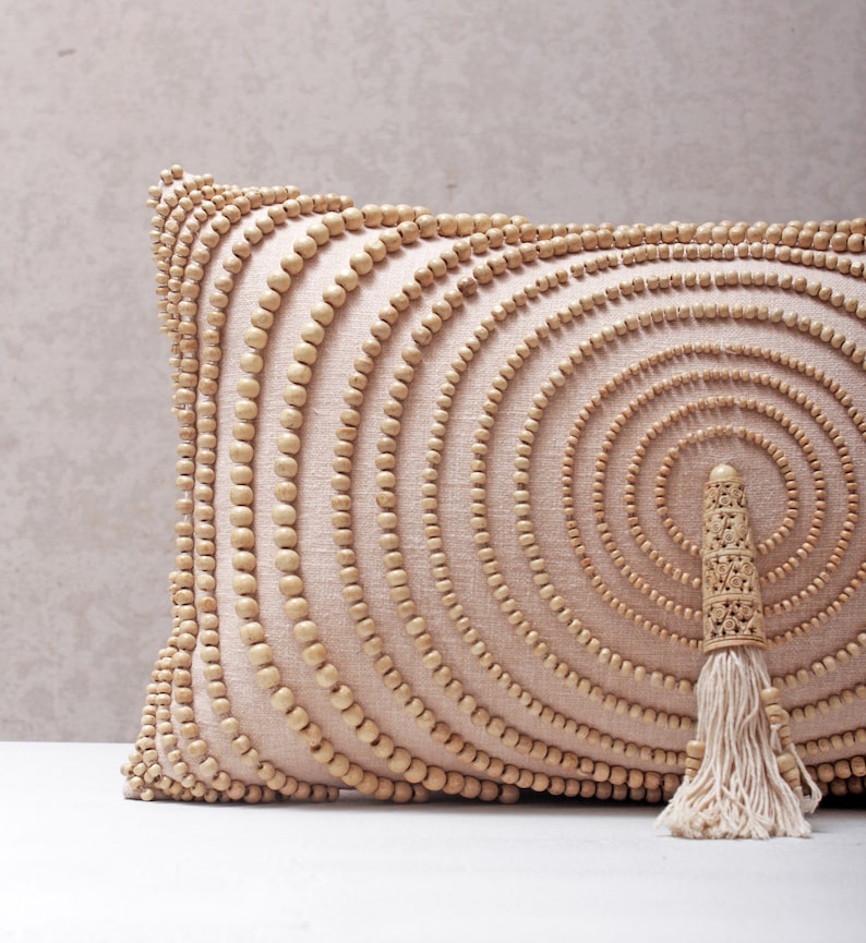 Puede incluir: Una almohada beige con un patr&oacute;n circular de cuentas de madera. La almohada tiene un borla de hilo blanco.