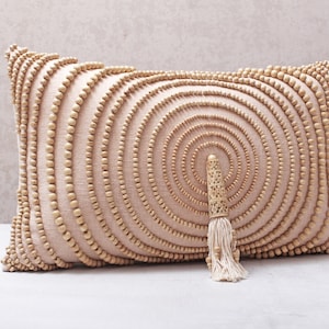 Puede incluir: Una almohada beige con un patr&oacute;n circular de cuentas de madera. La almohada tiene un borla hecha de fibras naturales.