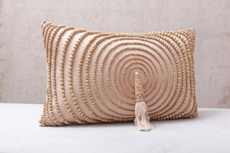 Puede incluir: Una almohada beige con un patr&oacute;n circular de cuentas de madera. La almohada tiene un borla hecha de fibras naturales.