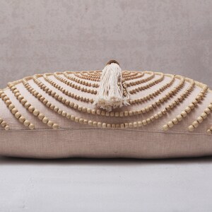 Puede incluir: Una almohada beige con un patr&oacute;n decorativo de cuentas de madera en un dise&ntilde;o circular. La almohada tiene un borla blanca.