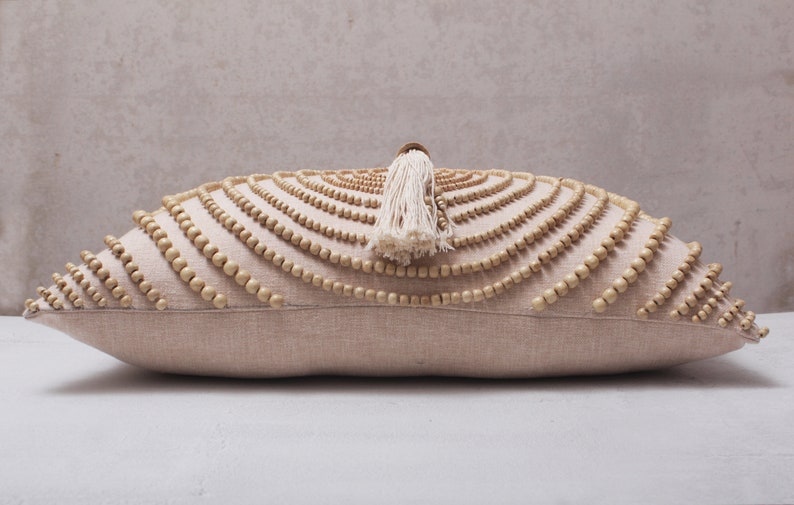 Puede incluir: Una almohada beige con un patr&oacute;n decorativo de cuentas de madera en un dise&ntilde;o circular. La almohada tiene un borla blanca.