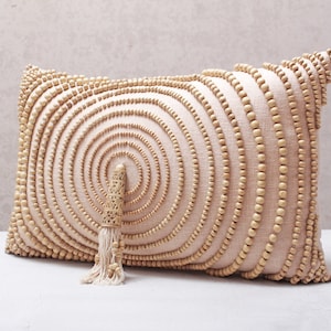 Puede incluir: Una almohada beige con un patr&oacute;n circular de cuentas de madera. La almohada tiene un borla hecha de fibras naturales.