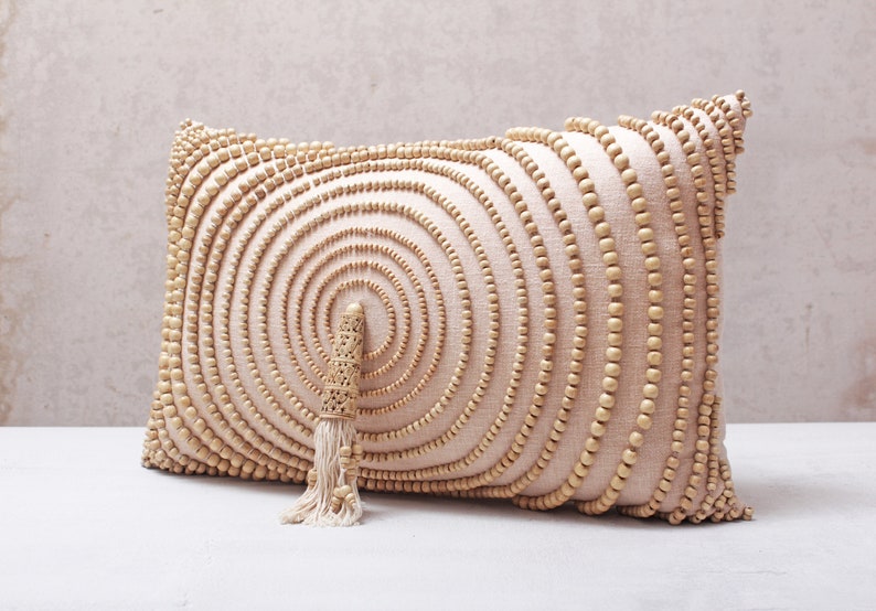 Puede incluir: Una almohada beige con un patr&oacute;n circular de cuentas de madera. La almohada tiene un borla hecha de fibras naturales.