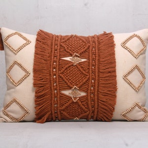 Puede incluir: Una almohada rectangular con una funda de algodón blanco y un patrón tejido marrón. La almohada tiene un borde de flecos y detalles de cuentas de madera.