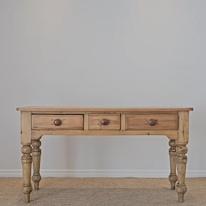 May include: A light-colored wooden console table with three drawers and turned legs. The table has a rectangular top and round wooden knobs on the drawers. The legs are thick and decorative, adding a rustic charm to the piece.