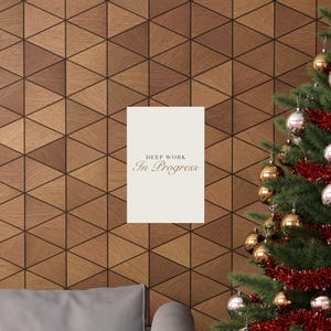 May include: A rectangular white wall sign featuring the black text DEEP WORK In Progress centered on the surface. The sign is positioned on a wall with a geometric wood triangle pattern next to a Christmas tree decorated with gold and silver ornaments.