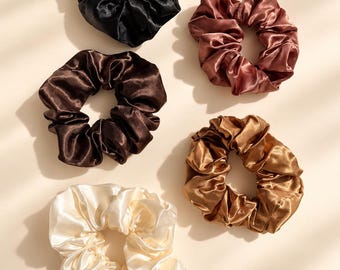 May include: A set of five circular hair scrunchies made of a smooth, shiny fabric. The collection includes one black, one dark brown, one cream, one copper, and one bronze scrunchie, each featuring a gathered, ruffled design with an elastic interior.