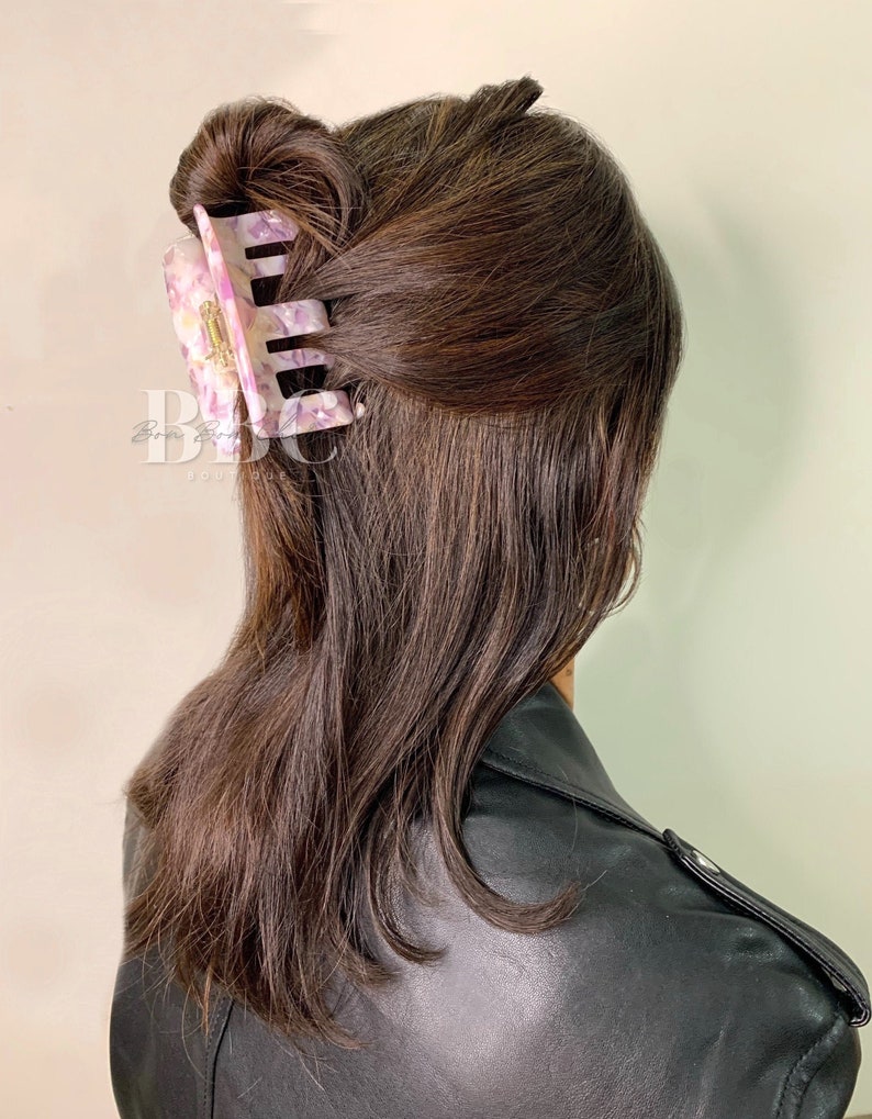 K&ouml;nnte beinhalten: Eine gro&szlig;e, rechteckige Haarspange in Rosa-, Wei&szlig;- und Lilat&ouml;nen, die einen Teil braunen Haares h&auml;lt. Die Spange hat ein marmoriertes Design und befindet sich am Hinterkopf. Die Person tr&auml;gt eine schwarze Lederjacke.