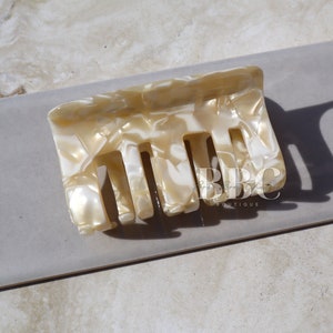 K&ouml;nnte beinhalten: Eine rechteckige Haarspange in einem marmorierten Muster aus Creme und Wei&szlig;. Die Spange hat eine Reihe von Z&auml;hnen zum Befestigen der Haare und besteht aus einem gl&auml;nzenden, harten Material. Die Spange wird auf einer neutralen, hellgrauen Oberfl&auml;che gezeigt.