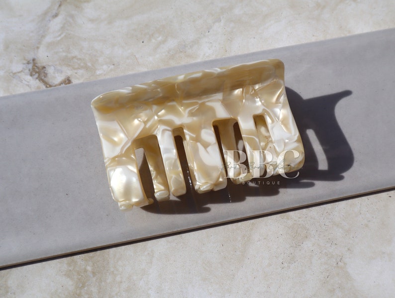 K&ouml;nnte beinhalten: Eine rechteckige Haarspange in einem marmorierten Muster aus Creme und Wei&szlig;. Die Spange hat eine Reihe von Z&auml;hnen zum Befestigen der Haare und besteht aus einem gl&auml;nzenden, harten Material. Die Spange wird auf einer neutralen, hellgrauen Oberfl&auml;che gezeigt.