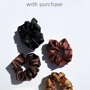 K&ouml;nnte beinhalten: Eine Auswahl an Satin-Haargummis in verschiedenen Farben, darunter Schwarz, Braun und Gold, wird auf wei&szlig;em Hintergrund pr&auml;sentiert. Der Text "FREE SCRUNCHIE with purchase" ist ebenfalls zu sehen. Diese Haaraccessoires sind ideal, um jedem Haarschnitt einen Hauch von Eleganz zu verleihen.