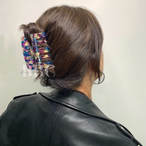 K&ouml;nnte beinhalten: Eine gro&szlig;e, farbenfrohe Haarspange in Blau-, Rosa- und Goldt&ouml;nen h&auml;lt einen Teil der braunen Haare zusammen. Die Spange hat eine rechteckige Form mit einem Schildpattmuster. Die Person tr&auml;gt eine schwarze Lederjacke.