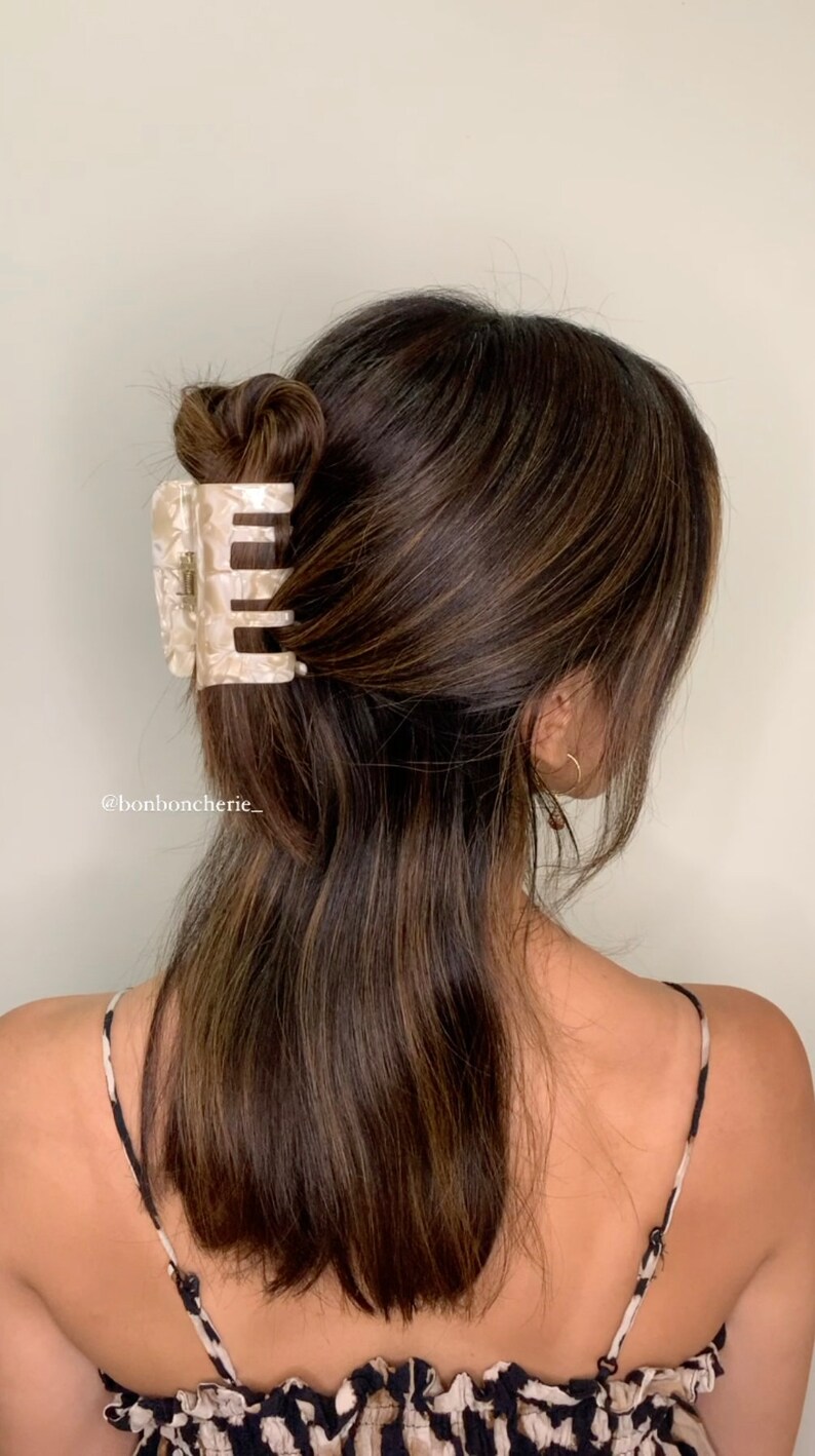 K&ouml;nnte beinhalten: Ein hellfarbener, gemusterter Haarspangenclip sichert einen Dutt in dunkelbraunem Haar. Der Haarclip ist rechteckig mit abgerundeten Kanten und einem perlmuttartigen Finish. Das Haar ist mit Mittelscheitel gestylt und f&auml;llt &uuml;ber den R&uuml;cken.