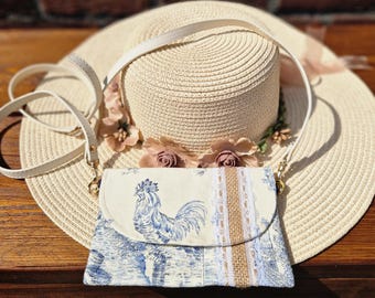 May include: A straw sun hat with a white strap and a floral accent sits on a wooden surface. A small, cream-colored clutch with a blue rooster design and a white strap is in front of the hat. The clutch has a burlap and lace stripe.