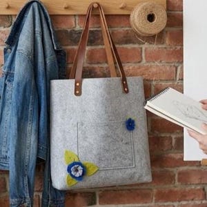 Może przedstawiać: Jasnoszara torba filcowa z dwoma brązowymi sk&oacute;rzanymi paskami na ramię przymocowanymi metalowymi nitami. Torba posiada dużą przednią kieszeń ozdobioną niebieskim kwiatem z tkaniny z zielonymi filcowymi liśćmi oraz małym niebieskim kwiatkiem.